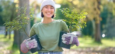 Gärtnern Eine Frau in Gartenkleidung hält Pflanzen in Töpfen in der Hand. Sie steht in einem Park mit Bäumen im Hintergrund
