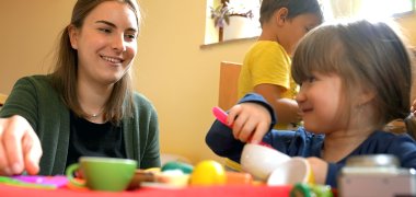 PivA Erzieherin (links) spielt mit zwei Kindern (rechts) in einer Spieleküche. Im Hintergrund ist eine KiTa-Inneneinrichtung zu sehen.