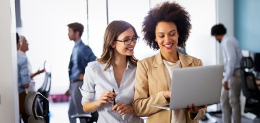 Verwaltungsfachangestellte Zwei stehende Frauen halten einen Laptop in der Hand und sehen lächelnd drauf. Im Hintergrund ist ein Büro zu sehen.