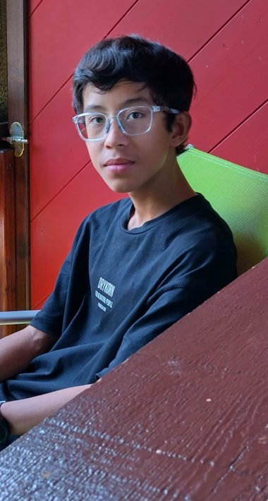 Louis Reckwerth Junger Mensch mit kurzen dunklen Haaren und klarer Brille sitzt auf grünem Stuhl vor roter Holzwand