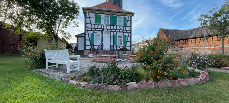 Rektor-Geißler-Haus mit Garten Rektor-Geißler-Haus in Dudenhofen, ein historisches Fachwerkhaus mit grün-weißen Fensterläden und einem Turm, umgeben von einem Garten mit Blumen und einer weißen Bank.