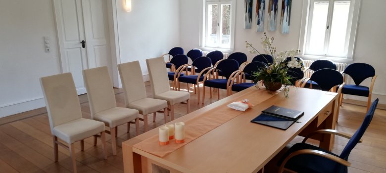 Trauzimmer im Rektor-Geissler-Haus Trausaal im Rektor-Geißler-Haus in Dudenhofen mit hellem Holztisch, vier beigen Stühlen, mehreren blauen Stühlen an den Wänden, zwei Fenstern und moderner Kunst an der Wand.