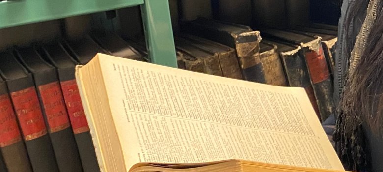 Archivnutzung Person hält aufgeschlagenes Buch vor Regal mit alten Archivbänden in Bibliothek und liest dieses.