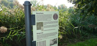 Informationstafel Informationstafel über Wasserburg und Blutgericht in einem grünen Park mit Büschen und Bäumen