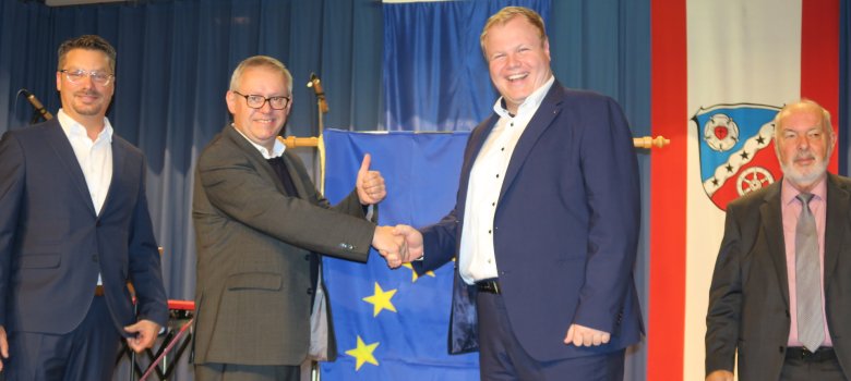 Verleihung der Ehrenfahne des Europarats Auf der Bühne stehen von links der ehemaliger Erster Stadtrat Schüssler, Pierre-Alain Fridez, Mitglied des Gremiums der parlamentarischen Versammlung des Europarates, Bürgermeister Breitenbach sowie der Stadtverordnetenvorsitzende Kaiser, im Hintergrund die Ehrenfahne sowie die Fahne mit dem Wappen Rodgaus.
