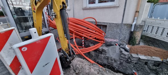 Glasfaserausbau in Rodgau Ein Bagger steht vor einem Haus, an dem der Bürgersteig aufgegraben ist und ein Glasfaserkabel verlegt ist.