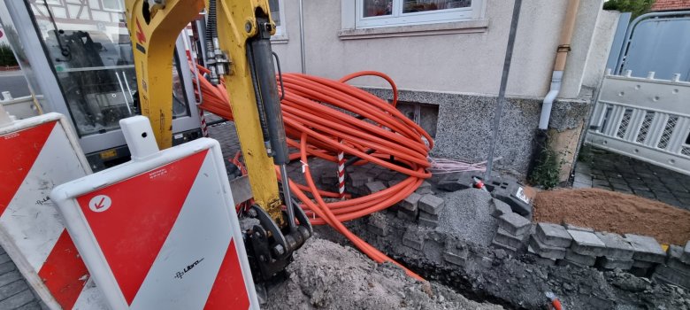 Glasfaserausbau in Rodgau Ein Bagger steht vor einem Haus, an dem der Bürgersteig aufgegraben ist und ein Glasfaserkabel verlegt ist.