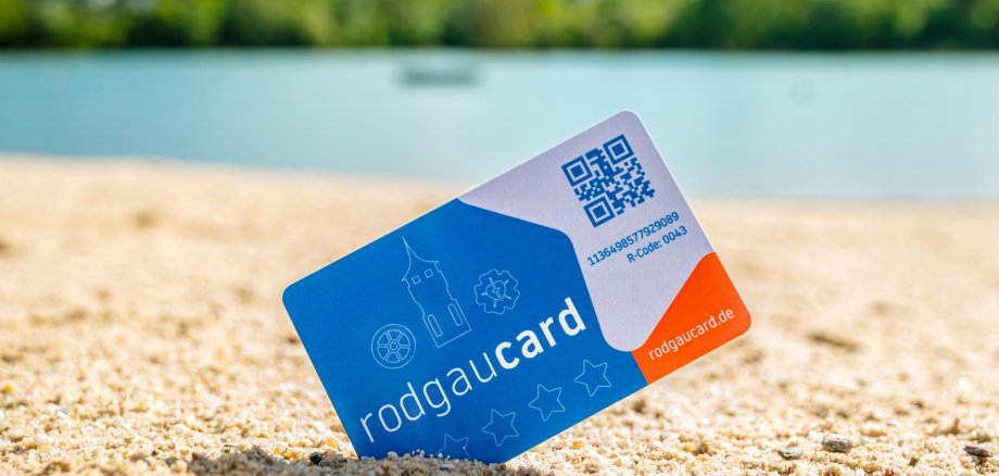 rodgaucard am Strand Rodgaucard-Karte steckt schräg im Sand am Strandbad mit unscharfem Wasser und grüner Ufervegetation im Hintergrund