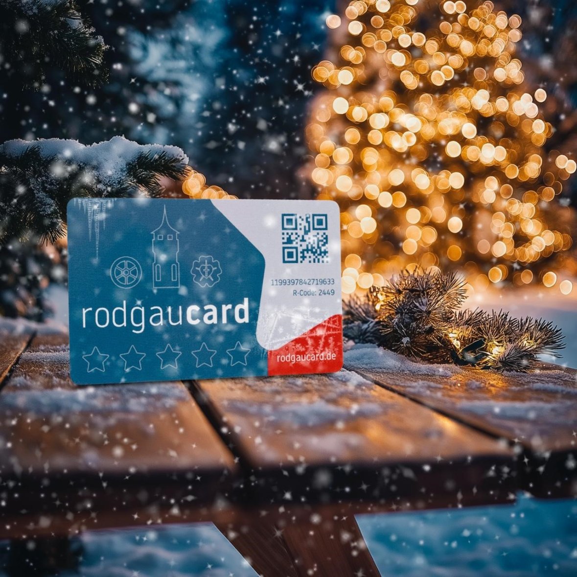 Winterliche rodgaucard rodgaucard in Blau mit weißen Symbolen und QR-Code liegt auf schneebedecktem Holztisch, im Hintergrund unscharfer, beleuchteter Weihnachtsbaum und Tannenzweige mit Schnee.