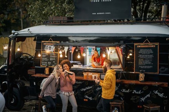 Symbol Reisegewerbe Reisegewerbe in Form eines Food Trucks mit beleuchtetem Verkaufsfenster, drei Personen bestellen und unterhalten sich im Freien.