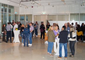 Viele Menschen stehen um hohe Tische in einem großen Geschäftsraum und unterhalten sich angeregt.