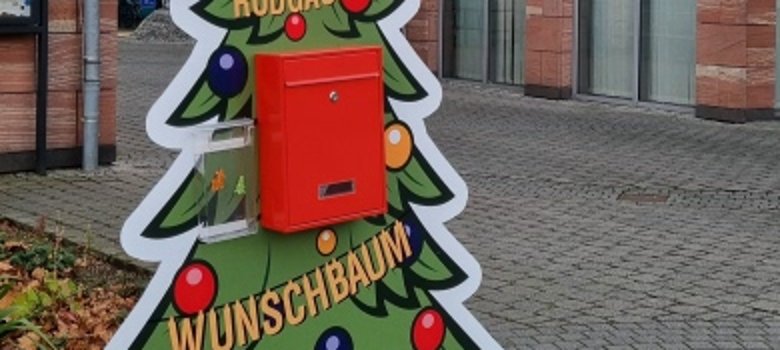 Wunschbaum am Rathaus Bunte Tafel in Form eines Weihnachtsbaums mit rotem Briefkasten, Aufschrift 'RODGAUER WUNSCHBAUM', steht auf gepflastertem Platz vor Rathausgebäude.