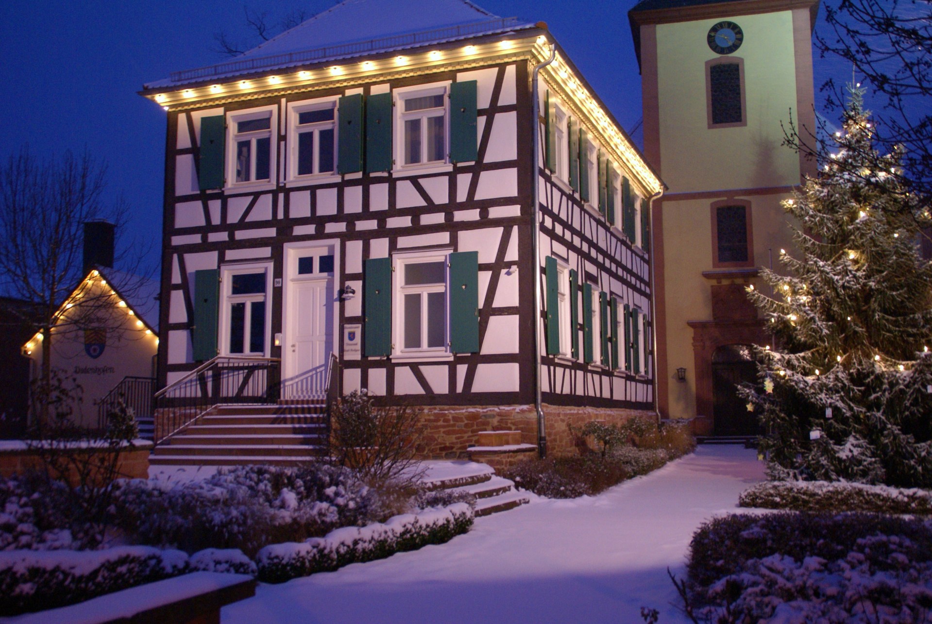 Rektor-Geißler-Haus im Winter Rektor-Geißler-Haus mit grünen Fensterläden und Weihnachtsbeleuchtung, davor schneebedeckter Garten und beleuchteter Weihnachtsbaum, im Hintergrund Kirchturm bei Abenddämmerung.